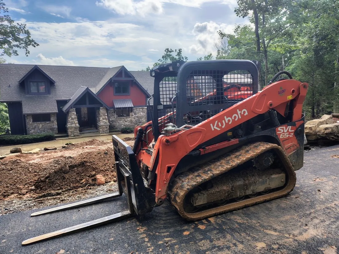 A Kubota compact track loader with pallet forks on a new drive in front of a stone and timber home, beside a pile of freshly graded dirt