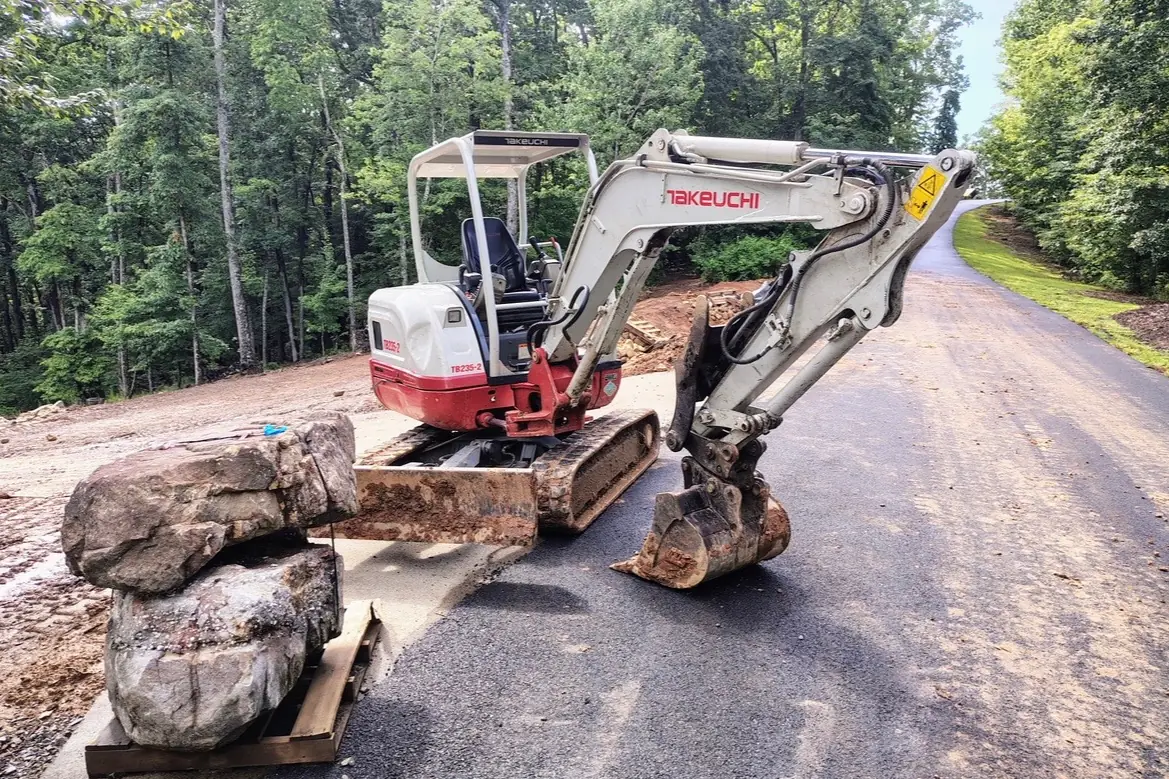 A Takeuchi mini excavator parked beside a newly paved drive, with a cleared and graded lot behind it and boulders staged on a pallet